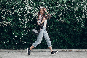 happy hipster woman with sunglasses having fun at blooming bush with white flowers of spirea. boho girl jumping and smiling in modern clothes, emotional moment. space for text