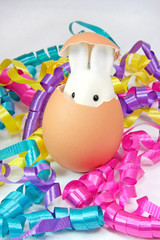 Cute Easter bunny peeking out from brown eggshell among a jumble of yellow, pink, blue and purple curled ribbons. Vertical. 