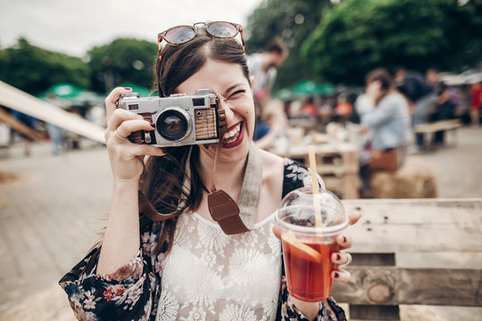 happy hipster woman in sunglasses making photo with old camera and drinking lemonade. stylish boho girl holding cocktail and smiling at street food festival. summertime