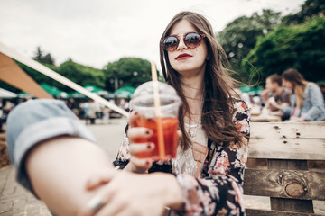 Fototapeta na wymiar stylish hipster woman in sunglasses with red lips holding lemonade. cool boho girl holding cocktail and sitting at street food festival. summertime. summer vacation travel. space for text