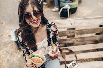 stylish hipster woman holding juicy burger and lemonade. boho girl holding  cheeseburger and smiling at street food festival. summertime. summer vacation travel. space for text