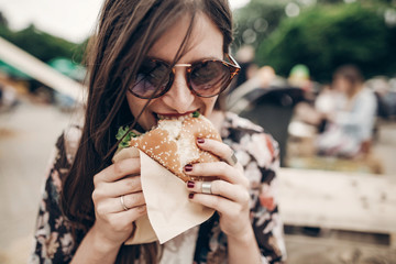 stylish hipster woman eating juicy burger. boho girl biting yummy cheeseburger, smiling at street food festival. summertime. summer vacation travel picnic. space for text - obrazy, fototapety, plakaty