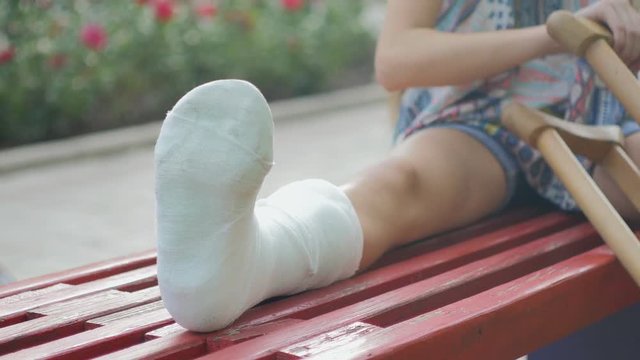 Girl with a broken leg sits on a bench, on the background of a descriptive pad and looks at the smartphone