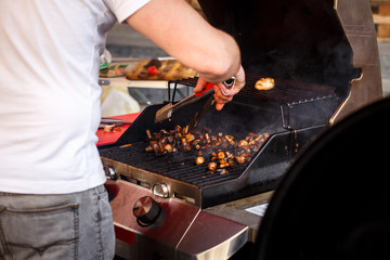 man roasting mushrooms, holding tongs, on open grill, outdoor kitchen. chef making vegetable meal, food festival in city. tasty food, food-court. summer picnic
