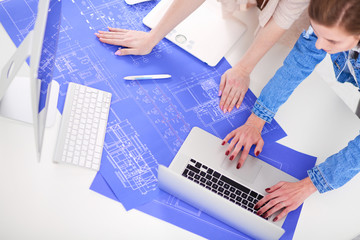 Two young woman standing near desk with instruments, plan and laptop.