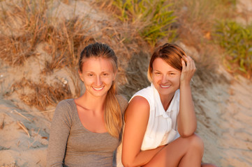 Fototapeta premium Two beautiful women sitting on sand at the beach look at the sunset