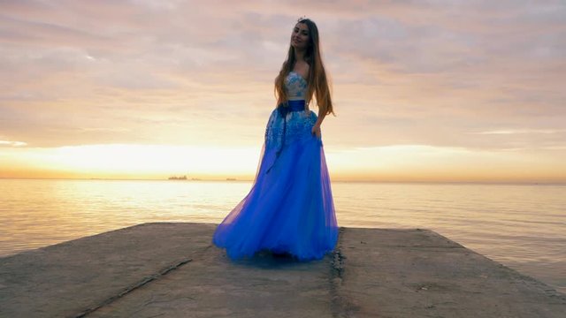 The young beautiful girl dances in a blue evening or ball dress on the pier close to sea. Slow motion.