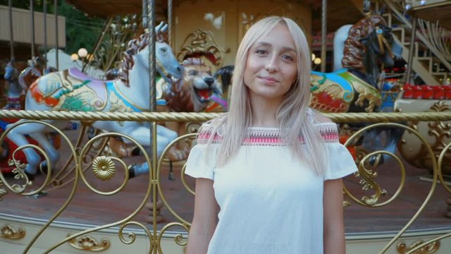 Young attractive blonde girl standing near carousel in amusement park. Beautiful portrait of woman with long hair. Slow motion.