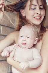 Young mother with her little blue-eyed baby wearing nothing relaxing and playing in the bedroom at the weekend together, lazy morning, warm and cozy scene. Selective focus. Brick wall background.