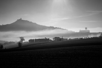 A view of a silhouette of St.Francis church in Assisi in the middle of mist, with cultivated fields in the foreground