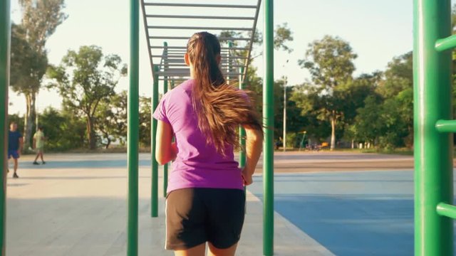Girl runs between horizontal bar in the morning. Sporty young attractive brunette woman making exercises. Slow motion. Back view.