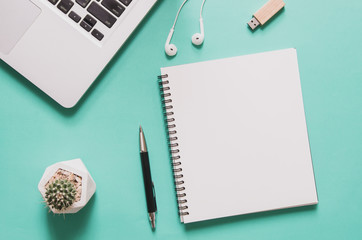 Office workplace concept. Computer laptop with blank notebook, cactus, pen, flash drive, earphone on blue background.