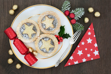 Christmas mince pie tarts on a plate with sliver fork, napkin and cracker with bauble decorations and foil wrapped chocolates on oak background. 