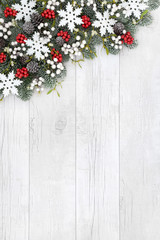 Christmas background border with white snowflake decorations, holly, mistletoe, ivy, fir and pine cones on rustic white wood.