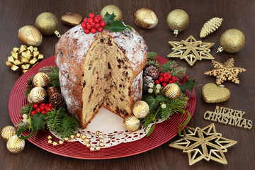 Chocolate panettone christmas cake with gold bauble decorations, holly, mistletoe, ivy, fir, pine cones on a red plate on oak wood background.