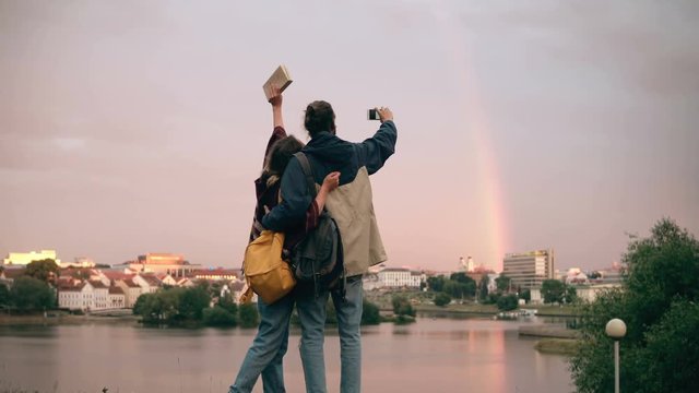 Back view of young happy couple taking photos of shore of the river and rainbow on smartphone. Man and woman having fun.