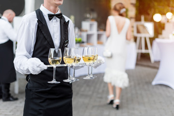 Midsection of professional waiter in uniform serving wine during buffet catering party, festive event or wedding. Full glasses on tray.