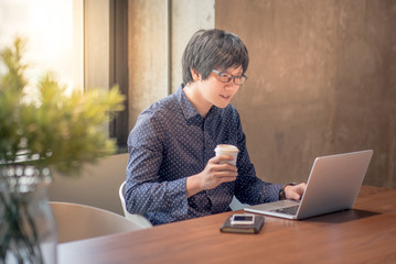 Young Asian businessman working with his laptop computer and holding coffee cup in office....