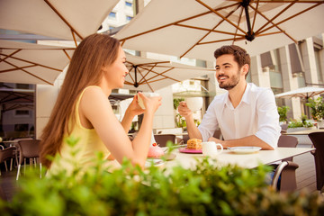 Young happy couple is sitting in light cafe`s terrace outdoors on a sunny day and enjoy, smiling, chatting with coffee and desserts © deagreez