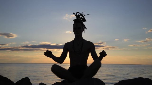 Silhouette of a man with native american indian feather mohawk accessory on head practicing yoga at the sunset.