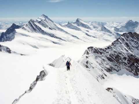 two mountain climbers on a narrow and exposed summit ridge descending from the summit with a spectacular panorama view of the Swiss Alps near the Aletsch glacier and Grindelwald