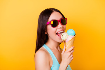 Cheerful young korean lady licks tasty ice cream of three scoops of different flavors, stands on yellow background in tourist wear, glasses