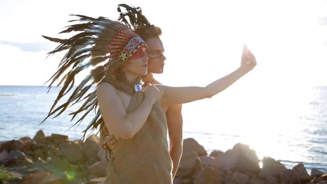 Young smiling Native American indian man and woman in Bohemian style sun wear dress taking selfie on sea beach at sunset.