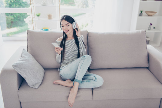 Excited beautiful asian teen is listening to music in big white earphones on her pda at home. She is wearing casual clothes, sitting on cozy beige couch
