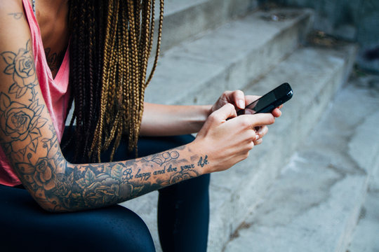 Hands of young woman with tattoos and braids using her phone sitting on stairs in a street