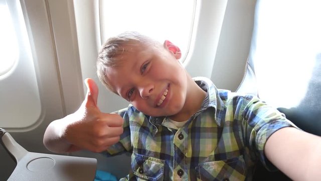 Closeup portrait of handsome happy funny little kid with thumb uo taking selfie with smartphone in airplane interior during flight. Point of view shot. Real time full hd video footage.