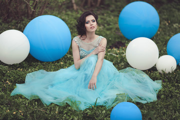 Happy girl in prom with helium air balloons. Beautiful girl graduate in a blue dress is sitting on the grass near a large balloons. © moredix