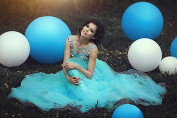 Happy girl in prom with helium air balloons..Beautiful girl graduate in a blue dress is sitting on the grass near a large blue and white balloons. Lovely girl with a beautiful smile. © moredix