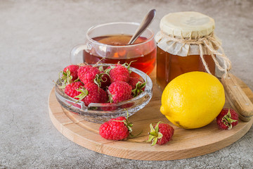  Prevention and treatment of influenza and colds. Ripe raspberries, fresh lemon, a glass jar with honey and a mug with hot tea on a round wooden board on a light background.
Used in folk medicine.