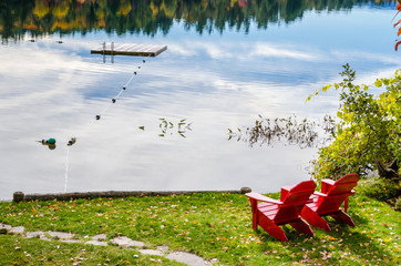 Two Red Adirondack Chairs on the Shore of a Lake and Reflection in Water