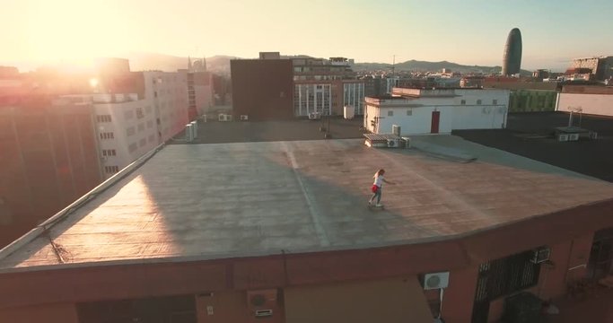 Young millenial teenager girl riding her surfskate on big roof top of big industrial building at sunset evening time, aerial shot with peaceful mood