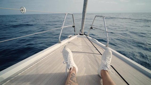 POV shot on two legs with white sneakers of relaxed male sitting on nose of sailing boat that going forward in wavy ocean or sea at summer evening time. Sxenic and peaceful b roll