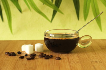 une tasse de café noir posé sur un sol en bois de bambou marron avec des morceaux de sucre et des grains de café sur fond vert à feuillage
