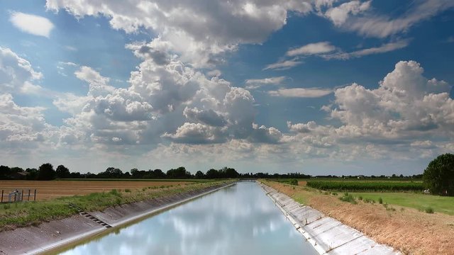 timelapse shot of clouds over irrigation channel, water flowing to supply cultivated fields in Italian countryside in color graded clip