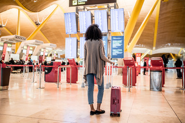 Black Woman looking at the timetable information panel in the airport with a suitcase