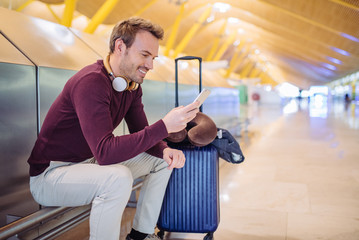 Young man waiting listening music and using mobile phone at the airport with a suitcase.