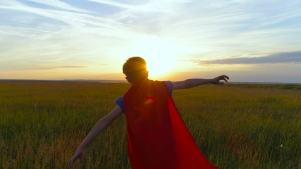 A boy in a superman costume runs across the green field at sunset
