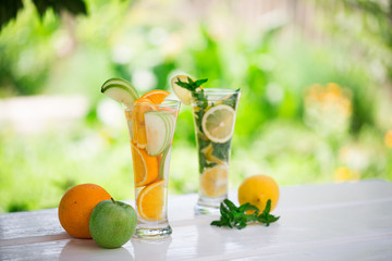 Two cocktail glasses of non-alcoholic mojito and detox fruit water orange-green apple and lemon-mint leaves on  white wooden table outdoors with garden background