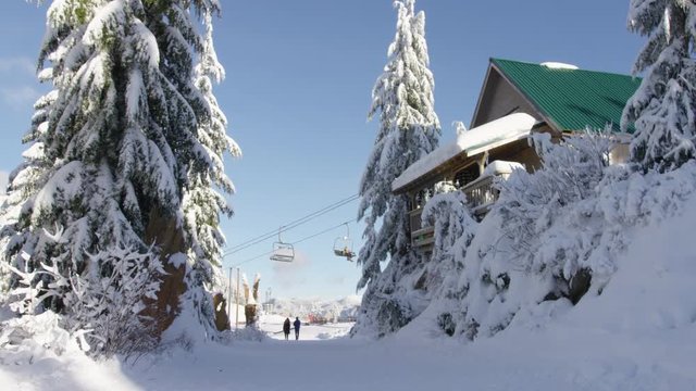 A shot moving down a snowy lane on Grouse Mountain with a cabin, chairlift and couple walking in the distance. 4k. Slow motion.