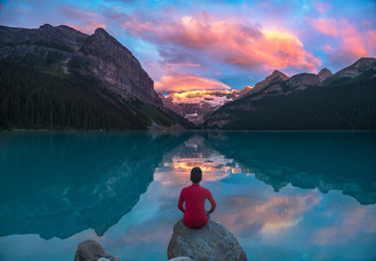 Man in red sit on rock watching Lake Louise morning clouds with reflections - obrazy, fototapety, plakaty