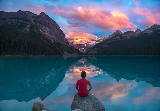 Man in red sit on rock watching Lake Louise morning clouds with reflections