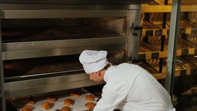 Young baker man in the uniform taking beatiful fresh croissant from oven and showwing it to the camera.