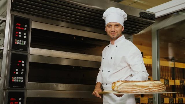Young baker man in the uniform taking beatiful fresh golden bread from oven and showing it to the camera.