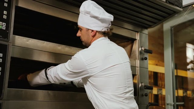 Portrait of young man in the uniform taking breads from oven, turning to the camer and possing.