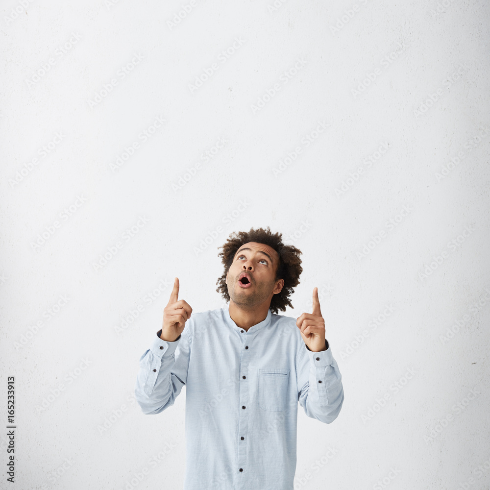 Wall mural Attractive dark-skinned male looking surprised or shocked upwards pointing his finger at white studio wall with copy space for your advertising content or promotional text. Astonished man advertising