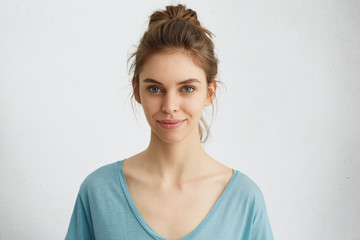Horizontal portrait of attractive blue-eyed female dressed casually having delightful look while smiling. Beautiful woman with hair bun having cheerful expression while posing at studio over white